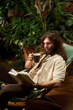 Vertical shot of Caucasian young adult man reading book while holding coffee cup indoors. Image ideal for mental wellbeing campaigns, lifestyle advertising and leisure concepts