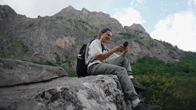 Young female traveler with a backpack using a smartphone while sitting on a large rock with a scenic mountain view during a hiking adventure.