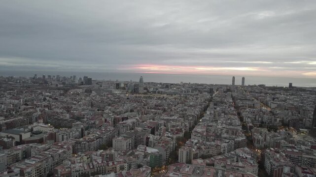 Aerial drone slow forward flight over Barcelona Eixample rooftops at dusk with orange glow on the horizon and twin towers by the sea, Spain
