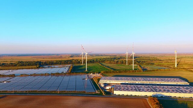 Solar farm and wind turbines, aerial view beside industrial buildings. Blue panel rows and warehouse roofs occupy the foreground beneath a cloudless sky.
