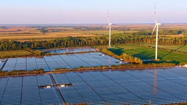 Solar arrays and two wind turbines, aerial view of neighboring energy sites. White towers rise above green plots behind blue panel rows in low directional sunlight.
