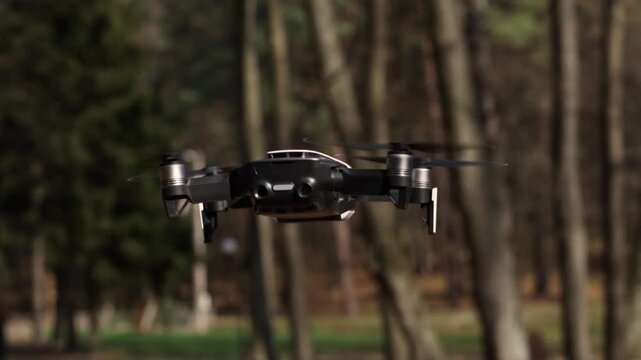 Black quadcopter drone hovering and flying away in a forest during an autumn day with spinning propellers