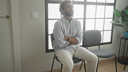 Fototapeta premium Man sitting on chair with arms crossed and hands clasped over stomach in building waiting area near window and plant; anxiety waiting.