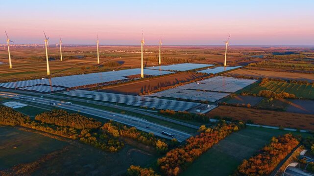 Solar arrays and wind turbines, aerial view beside a divided highway. Blue panel blocks lie beyond traffic lanes and tree belts beneath a pink evening horizon.