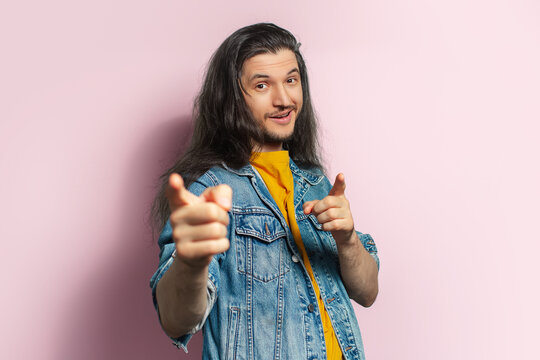 Young happy man with long hair pointing index finger on camera. Isolated on pastel pink background.