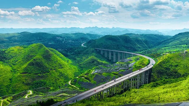 High elevated highway overpass curves through lush green karst mountains near solar panel farms in Guizhou under clear sky.