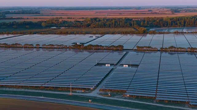 Photovoltaic power station in countryside at dusk. High angle shot displays rows of solar modules next to road and trees.