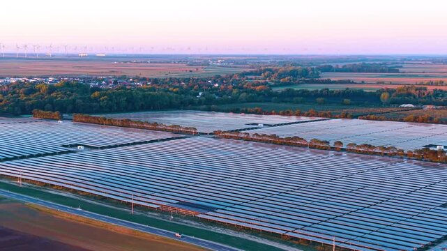 Solar farm, aerial view toward a village and cultivated fields. Blue panel rows extend past a road and tree belts toward clustered houses under a pink sky.