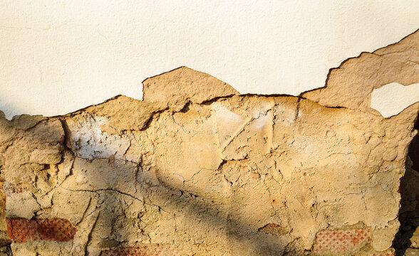 Texture background with crumbling beige plaster on a weathered wall and exposed red brick edges surrounded by deep cracks and worn surface