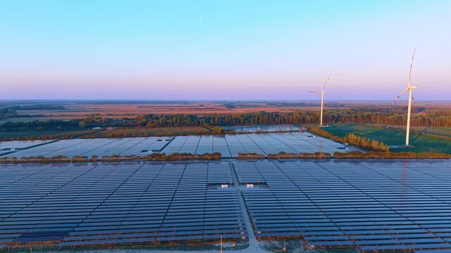 Photovoltaic park with wind turbines in background. Drone view shows blue solar rows extending toward distant wind turbines.