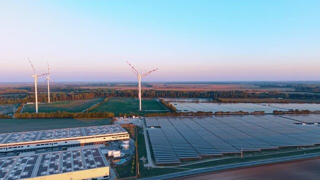 Solar farm and wind turbines, aerial view beside warehouse rooftops. Industrial buildings border blue panel blocks below white towers under a clear evening sky.