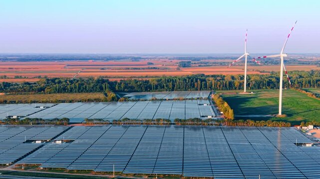 Solar array blocks and wind turbines, aerial view of rural power infrastructure. Blue modules form parallel bands below white towers, tree belts and cultivated plots in low sunlight.