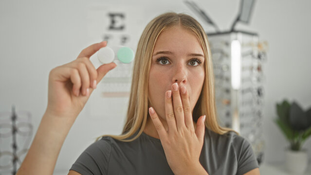Young woman holding a contact lens case up, hand to lips covering mouth, eyeglasses display and eye chart visible in building optician office; surprise uncertainty.