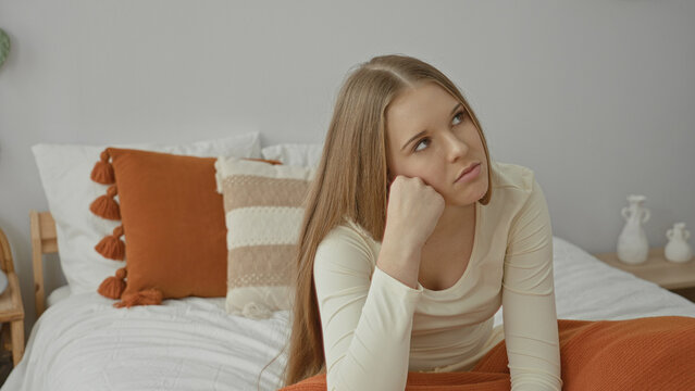 Young woman rests head on fist on bed with orange blanket and tassel pillow, then rubs forehead and leans against headboard in bedroom; fatigue contemplation.
