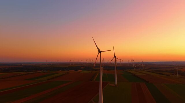 Wind farm, aerial view of two foreground turbines over cultivated plots. Striped green and brown fields extend toward distant towers beneath an orange evening horizon.