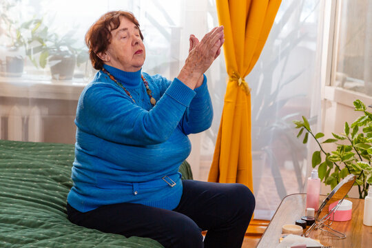 Elderly Caucasian woman sitting comfortably on a bed while caring for facial skin with cream. The scene represents beauty, wellness, personal grooming, confidence, and positive aging. Mid shot
