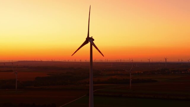 Single wind turbine silhouette against bright yellow dusk. Dark mechanical blades stand centered over countryside terrain in warm evening light.