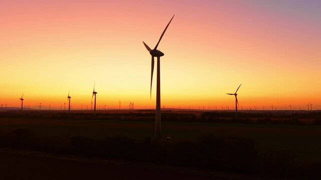 Wind turbine silhouette, foreground rotor against a sunset sky. A tall tower stands above dark fields, with smaller turbines beneath a yellow and orange horizon.