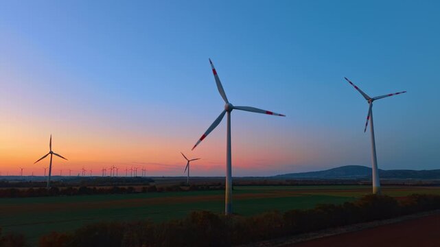 Wind turbines, aerial view over farmland in blue twilight. White towers with red blade tips stand above green plots and a road near a pink horizon.
