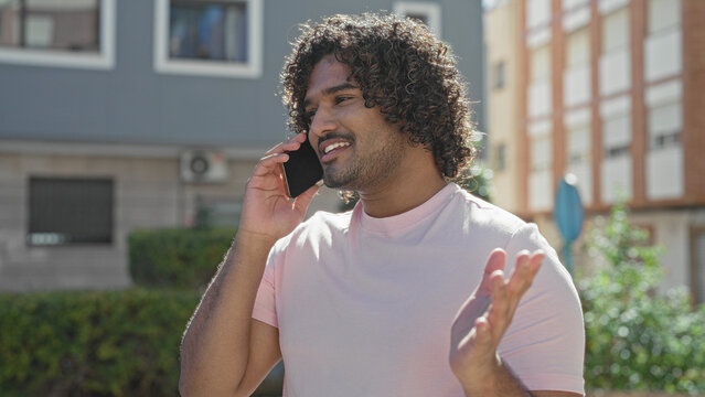 Man holding smartphone to ear, smiling and gesturing on a sunny street in front of buildings; friendly conversation cheerful.