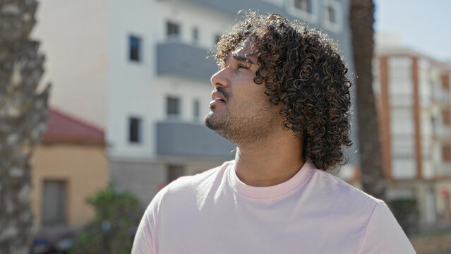 Man shielding eyes with hand on street by apartment building, squinting from bright sun; irritation.