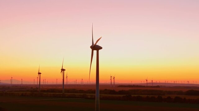 Wind turbines, backlit towers over fields beneath a pink sunset sky. A foreground rotor appears in side profile, with smaller towers spaced across the yellow horizon.