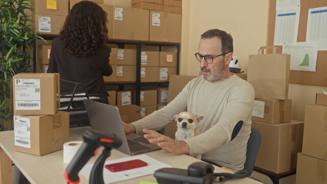 Man typing on laptop with small dog on lap and stacked shipping parcels, scanning labels with barcode reader in building; focused productivity.