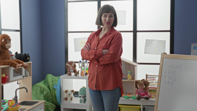 Woman teacher with arms crossed by a whiteboard, toys and children's drawings in a daycare room inside a building; nurturing calm.