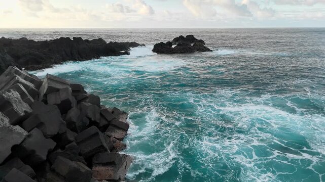 Turbulent ocean water beside concrete breakwater and volcanic reefs. Sea foam across coastal water surface