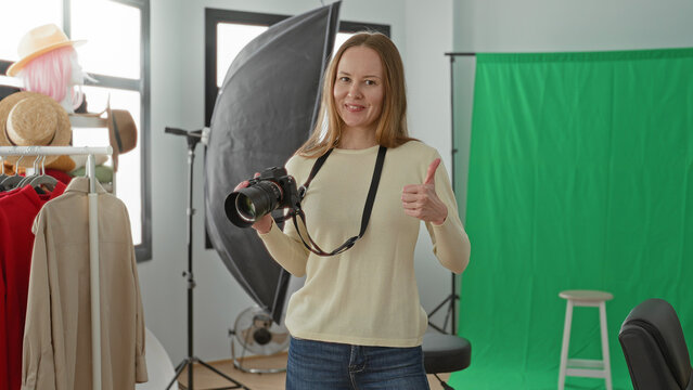 Young woman photographer holding dslr camera with strap and giving thumbs up amid green screen, softbox, mannequin head, clothing rack and stool in studio; confidence creativity.