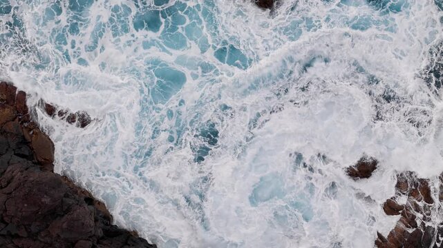 Breaking Atlantic ocean waves and sea foam between volcanic rocks. Overhead aerial view of turbulent water along rocky shoreline