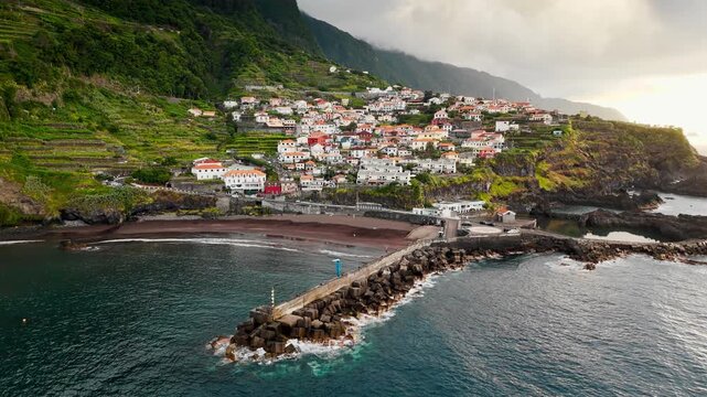 Seixal beach beneath coastal mountains in Madeira, Portugal, aerial view. Black sand shoreline along Atlantic bay beside village