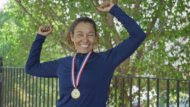 Woman wearing gold medal raises both arms and flexes fist, smiling broadly in front of leafy fence at forest; pride victory.