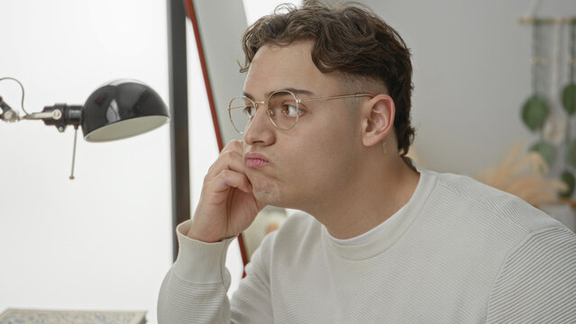 Man in white sweater wearing glasses, pouting and pressing cheek with hand while seated at a desk lamp in studio; quiet boredom.