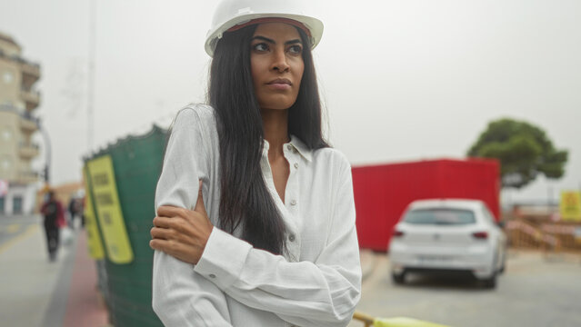 Woman architect wearing hardhat holds left arm across chest and grips elbow on street by construction site; professional determination.