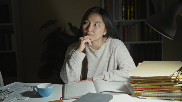 Woman with hand on chin studying at desk with open notebook, blue cup, lamp and tall stack of folders, bookshelf visible in a studio; contemplation.