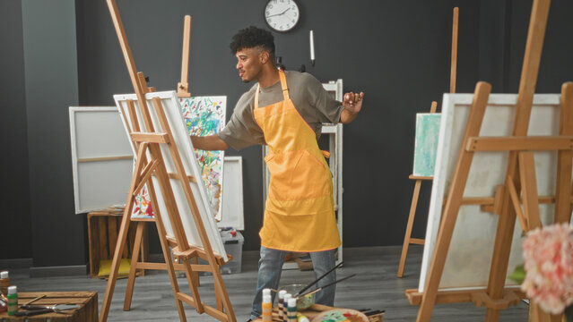 Man painter in yellow apron holds paintbrush in right hand while stepping and dancing beside canvas on studio easel; joyful creativity.