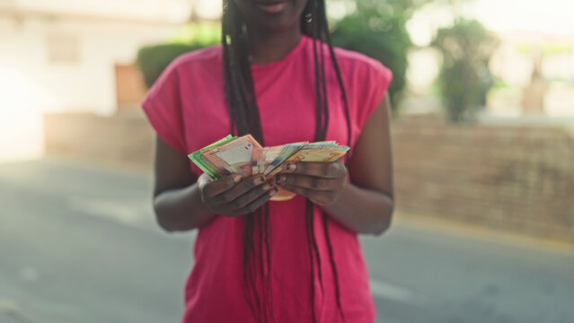 Young smiling woman counting rand banknotes with hands on a sunny residential street; joy saving independence.