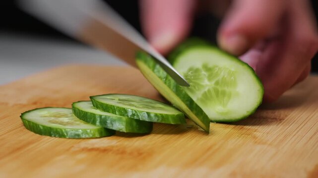Close-up of fresh cucumber being sliced with a kitchen knife on wooden cutting board. Healthy vegetable preparation and everyday cooking process in the kitchen.