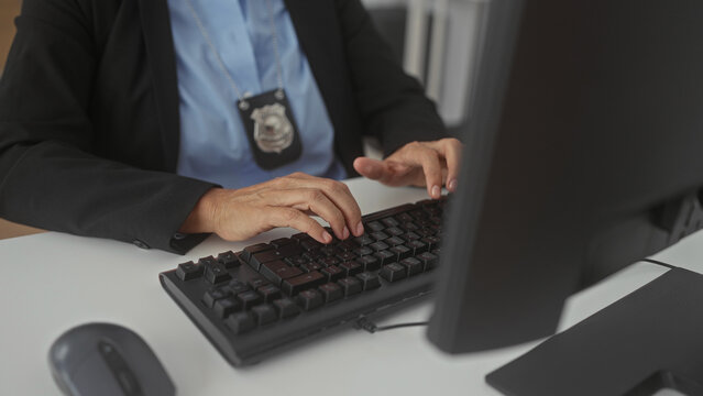 Woman police officer typing on mechanical keyboard at office desk with visible badge and computer monitor in building; focused investigation.