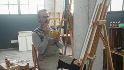 Man painter in apron sits at easel with palette, holding yellow coffee cup, hand on chin in studio  contemplative calm. © Krakenimages.com