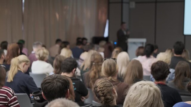 Conference attendees watch a speaker in a dark jacket as the camera pans right toward projected slides.