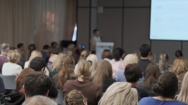 Seated attendees face projected slides as one participant raises a phone during rightward camera panning.