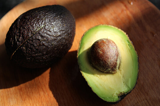 Fresh ripe Hass avocado halved with seed and a whole fruit on wooden cutting board in bright natural sunlight with deep shadows