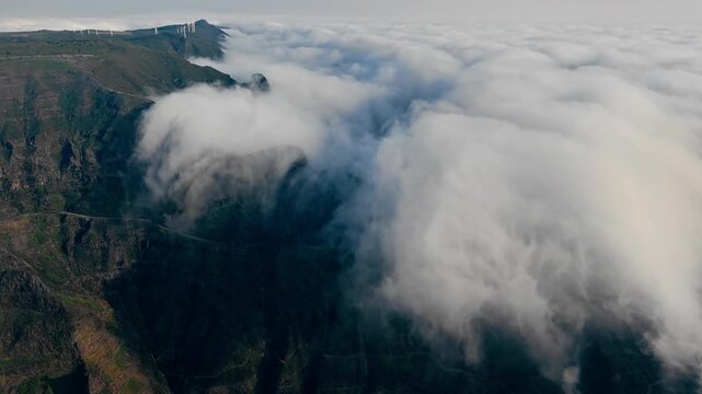 Dense clouds advancing into mountain valley and cascading down steep slopes. Ominous aerial landscape with cliffs disappearing beneath cloud cover