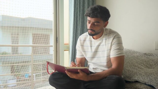 Young man sitting comfortably on a bed and writing in a personal journal at home. Suitable for journaling, self-reflection, personal development, studying, writing, mindfulness.