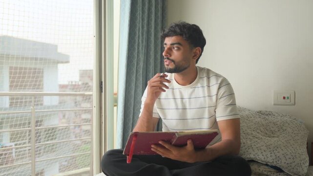 Young man sitting comfortably on a bed, writing in a journal and pausing to think about his thoughts and ideas. Natural window light creates a calm home environment.