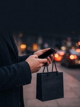 Person holding smartphone and black shopping bag on night city street