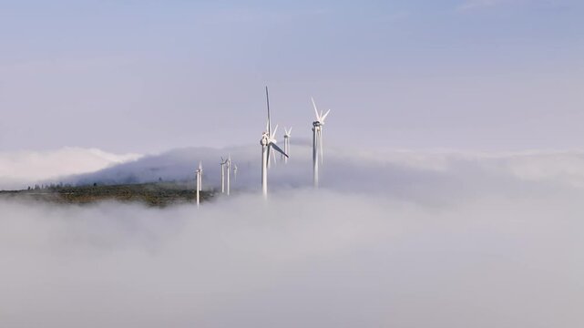 Wind energy infrastructure partially hidden by low clouds in mountainous terrain. Renewable electricity generation above cloud layer
