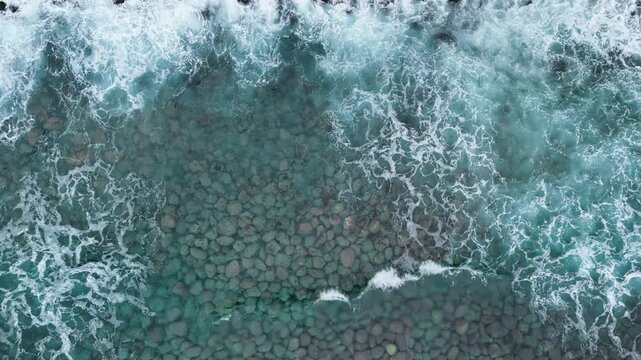 Top view drone shot of Atlantic waves washing across rounded rocks visible through clear water, detailed coastal texture